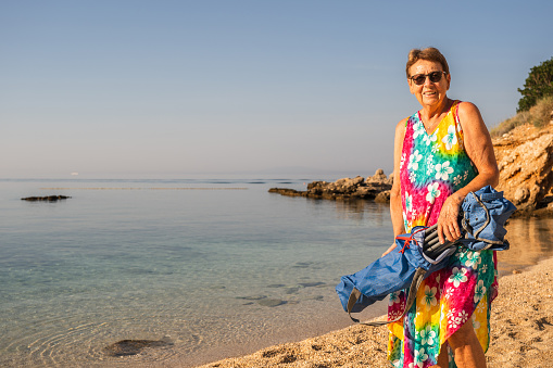 happy senior woman in colorful summer beach dress with camping chair on the beach on croatian island of krk, smiling at camera