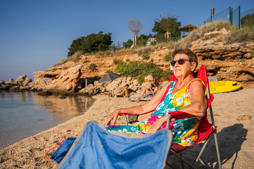 senior woman sitting in camping chair on a small beach with calm waters of adriatic sea on sunny summer morning
