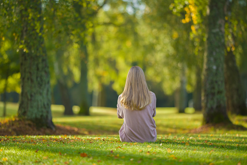Young adult blonde woman sitting on green grass at city park in beautiful warm sunny autumn day. Spending time alone and enjoying freedom at nature. Peaceful atmosphere. Back view.