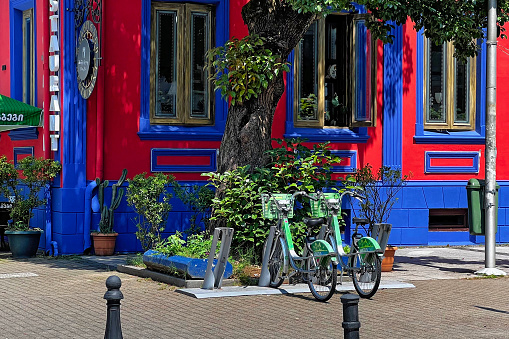 Batumi, Georgia - June 29, 2023: Green rental bicycles parked in front of a vibrant blue and red building with a tree in Batumi.