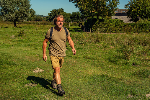 Man in green t-shirt and shorts with backpack walks in sunny countryside.