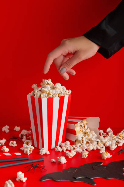 istock Womans hand taking popcorn from striped box, bat napkin and straws on a red background. 1718491784