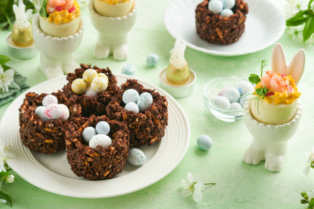 istock Easter chocolate nest cake with mini chocolate candy eggs with blossoming cherry or apple flowers on green background table. Creative recipe for Easter table with holiday decorations. Top view. 1718502476