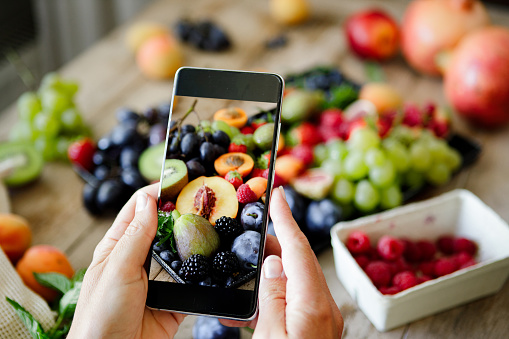 Close-up of woman photographing fruits on kitchen table with her mobile phone. Point of view of a woman hand holding a cellphone and taking photos of a variety of fruits on a tray over wooden table.
