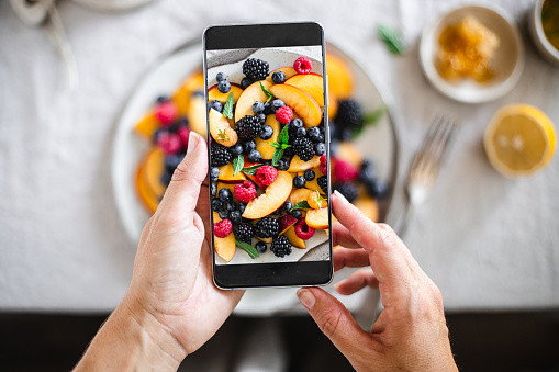 Close-up of a woman photographing freshly made fruit salad.  Female taking picture of healthy breakfast on table with her mobile phone.