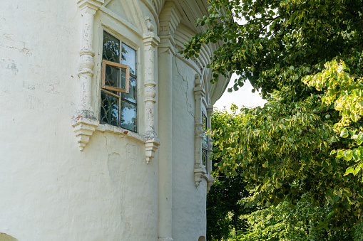 Fragment of the wall of an old church with small windows decorated with stucco details in the traditional Russian style, white plaster, windows to the garden, green vegetation, architectural background, summer.