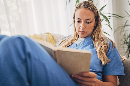 Female nurse came back from work and still wearing scrubs relaxing at home and reading a book