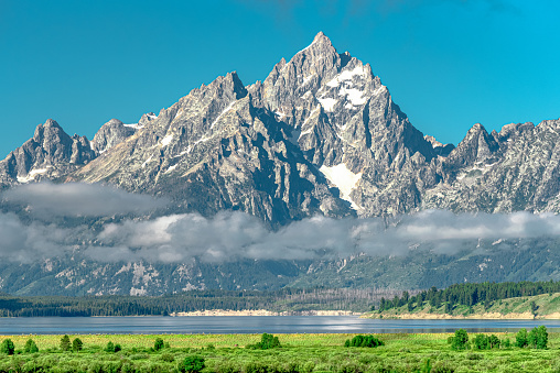 The snow-capped peaks of the Grand Teton mountains reach into the bright blue sky while a thin layer of fog rises above Jackson Lake in the morning hours at  Grand Teton National Park, Wyoming.