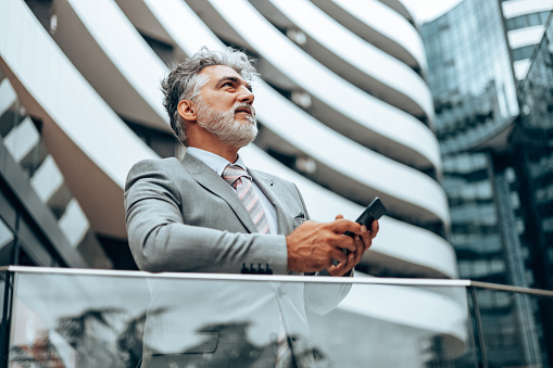 Handsome senior businessman standing at street and using smart phone for communication. Modern buildings in background. Low angle.