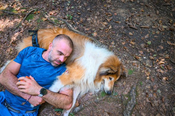 Adult man in blue sport clothes lying with his old injured dog on the ground in forest.