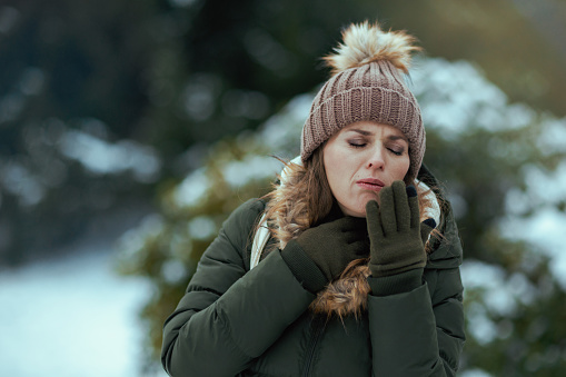 unhappy modern woman in green coat and brown hat outdoors in the city park in winter with mittens and beanie hat coughing.