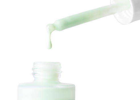 Drop falls from a pipette into a cosmetic bottle isolation on white background, macro. Pastel green face cream, makeup primer, color correcting cosmetic product, serum