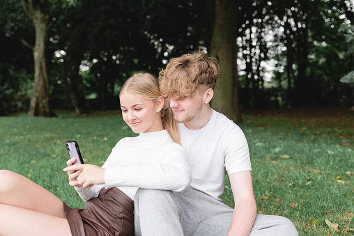 A young teenage couple sitting on the grass sharing a mobile phone