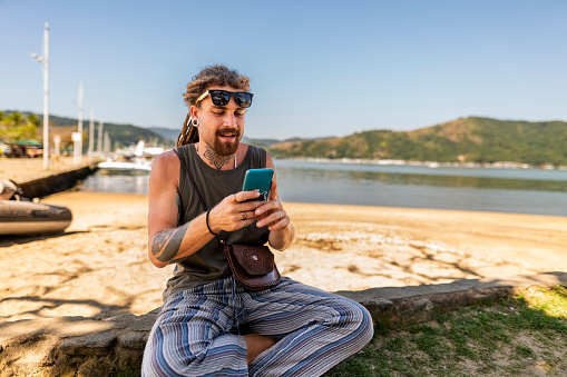 Handsome young man using mobile phone while sitting near a sea bay on summer day