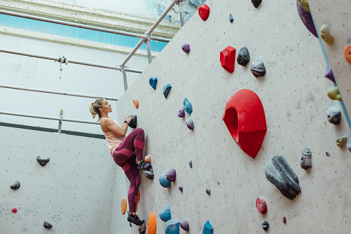 A low angle view of a mid adult woman who is setting herself new personal bests while enjoying a successful indoor climbing session at an indoor climbing centre in Newcastle upon Tyne in the North East of England. She is almost at the top of an intermediate level wall and is steps away from the top.