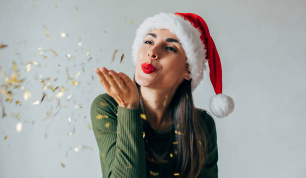 Close up photo of excited young cute woman in Christmas hat is blowing confetti from her hands and celebrating New Year on party stock photo