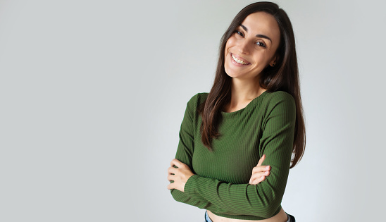 Portrait of young hispanic woman with toothy smile and crossed arms isolated on grey wall with copy space. Gorgeous girl in casual wear looking at camera against grey wall