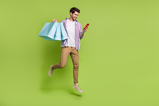 Full size photo of cheerful person flying hold shopping bags look at smartphone order new clothes isolated on green color background.