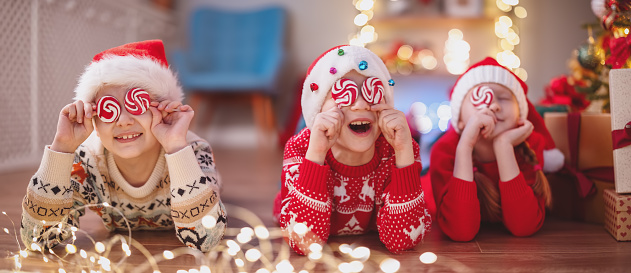 Smiling children lying on the floor indoors and covering by lollypops their eyes. Concept of the Christmas, New Year and family holidays together.