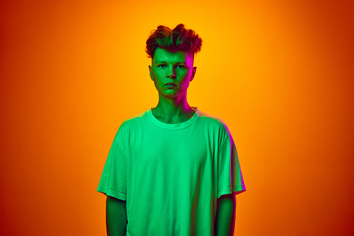 Portrait of calm young man, guy, student in white t-shirt isolated on gradient orange background illuminated green neon light. Concept of youth, beauty, fashion, trends emotions, sales. ad