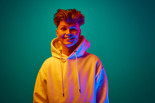 Portrait of funny young handsome guy, man, student dressed white hoodie looking at camera against gradient mint background in mixed colors neon filter. Concept of emotions, beauty, fashion, style. ad