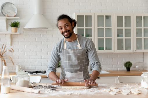 Head shot portrait of smiling African American man rolling out dough, standing in kitchen at home, happy young bearded male wearing apron cooking homemade cookies or pie, looking at camera
