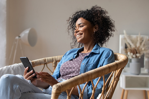 Dreamy happy African American woman holding smartphone, looking in distance, smiling attractive young female excited by good news, chatting online with boyfriend, relaxing sitting in armchair at home