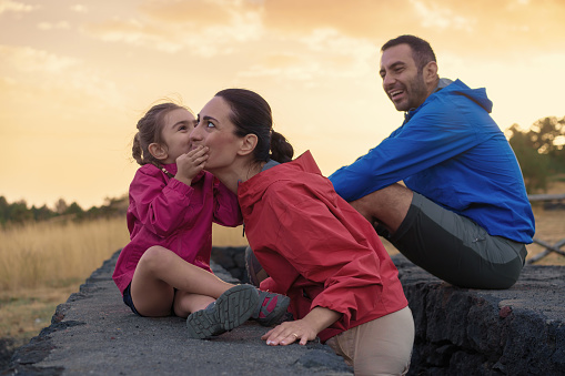 A family rests on a basalt stone wall after a mountain hike. A child sweetly kisses her mother's cheek, drawing amused smiles from both parents.