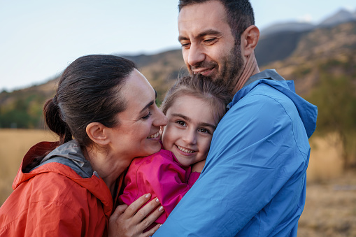 Mediterranean Family Moment in Mountains: A loving father and mother cuddle their attentive daughter during an outdoor mountain adventure, as she looks at the camera.