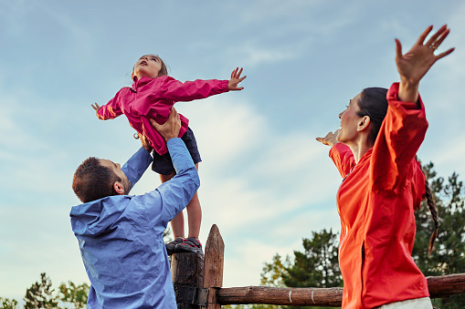 Under a vast blue sky, a playful daughter stands on a fence, imagining flight as her father supports her. With outstretched hands, she 'flies' while her mother demonstrates the wing movements.