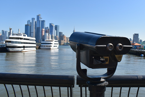 Coin-operated binoculars provide a close-up view of Manhattan's iconic skyscrapers and luxurious yachts along the waterfront, offering a stunning New York City landscape.