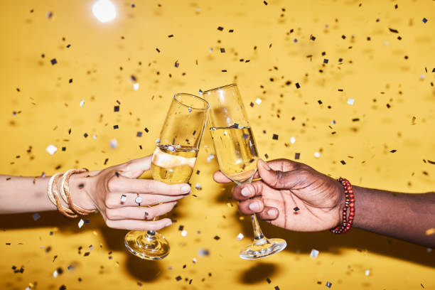 istock Two hands clinking champagne glasses against yellow background with confetti 1718782019