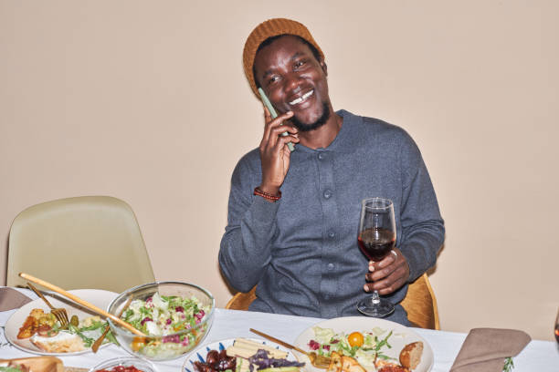 夕食の席でスマートフォンで話す笑顔の若い黒人男性 - dining table flash ストックフォトと画像