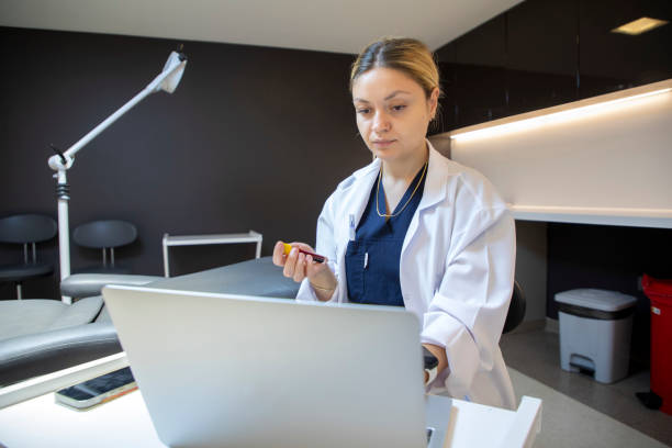 a young blonde medical staff member is holding a blood tube and typing information on her laptop in the clinic. - dentist office nurse doctors office blond hair imagens e fotografias de stock