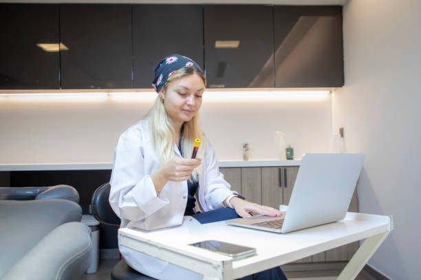 a young blonde medical staff member is holding a blood tube and typing information on her laptop in the clinic. - dentist office nurse doctors office blond hair imagens e fotografias de stock