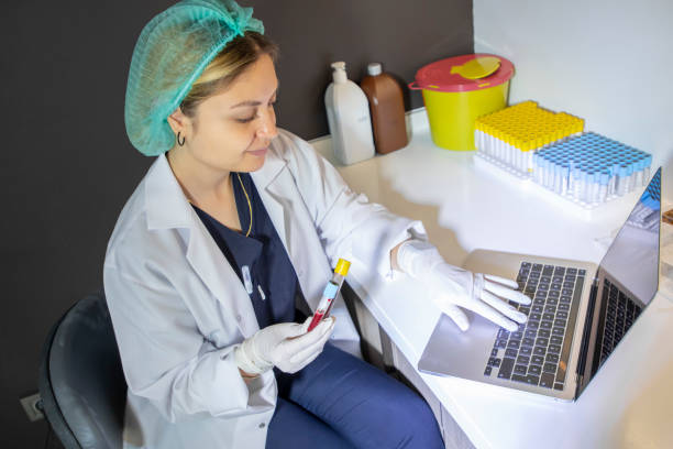 a young blonde medical staff member is holding a blood tube and typing information on her laptop in the clinic. - dentist office nurse doctors office blond hair imagens e fotografias de stock