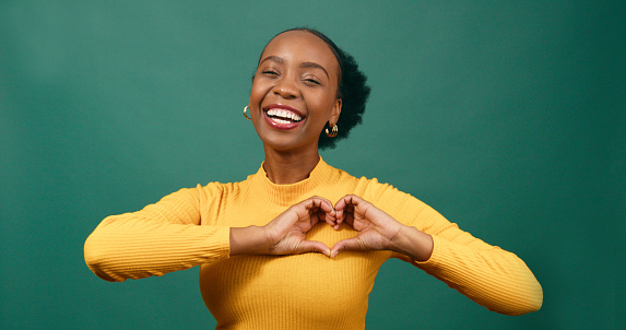 Young Black woman forms heart with hands to chest, smiling green studio. High quality photo
