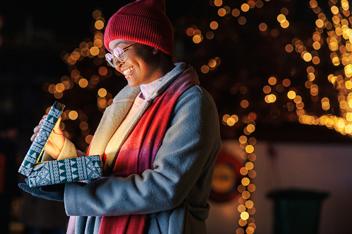 A young African American woman wearing winter clothing is standing outside and opening a Christmas present with a smile on her face. There is a light shining from the opened box.