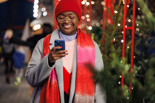 A young African American woman dressed in warm winter clothes cheerfully using her mobile phone. There's Christmas decoration behind her.
