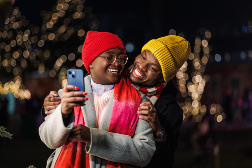 A young African American couple cheerfully embracing under the Christmas lights, while taking a selfie on a mobile phone. They're wearing warm winter clothing.