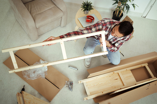 Young casually clothed man checking wooden piece of shelf while assembling furniture at home
