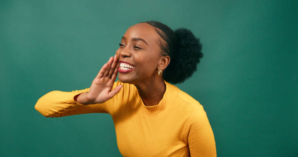 Young Black woman raises hand to mouth, yells excitedly, green background studio stock photo