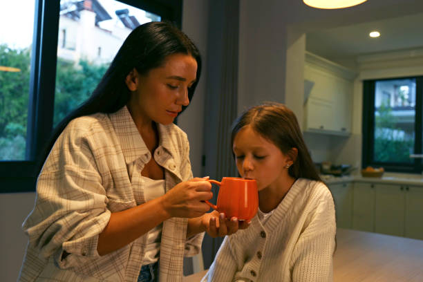 Mom giving her daughter a try of a tasty hot chocolate drink. Parental care concept. Mother and her six year old girl drinking from a pumpkin shaped cup. Close up, copy space, kitchen background.