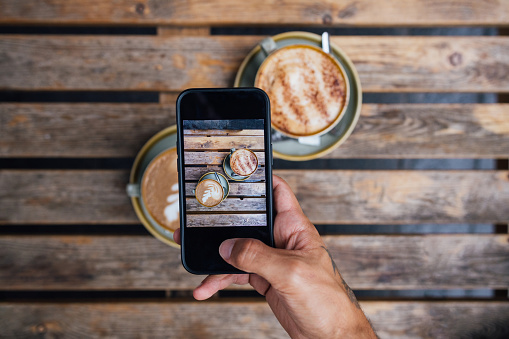 A directly above shot of two coffees with latte art on an outdoor picnic table. An unrecognisable person is taking a photo of the cups on his smart phone.