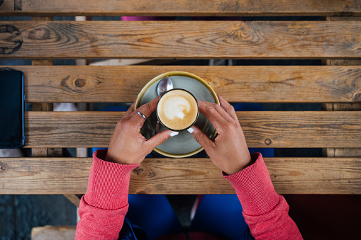 A directly above shot of an unrecognisable woman holding a coffee with latte art on an outdoor picnic table.