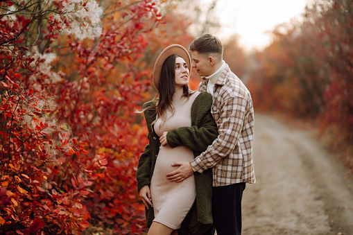 A man and his pregnant wife are walking in an autumn park, spending time together. Concept of love, relaxation, people. Active lifestyle.