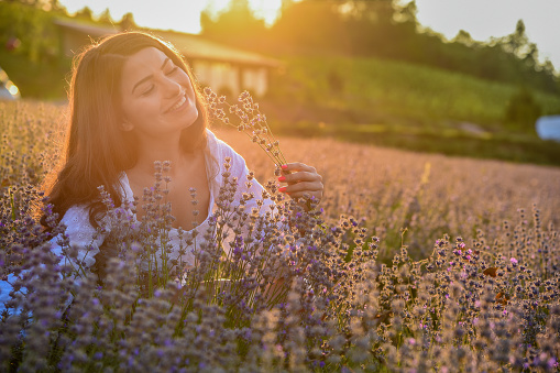 A girl with a wide smile on her face sits in a field of lavender and plays with a sprig of lavender as the sun sets