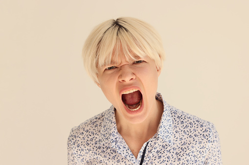 Expressive blonde woman screams with wide open mouth squinting eyes. Young female person with stylish short haircut shows emotion of anger playing role