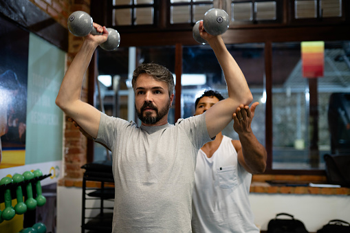 Mature man exercising with dumbbells during a strength training session at the gym with a trainer assisting him