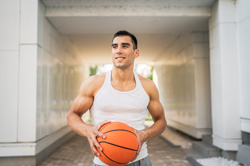 One young caucasian man male athlete stand outdoor hold basketball ball wear white tank top a-shirt strong muscular real person copy space happy smile confident healthy lifestyle concept